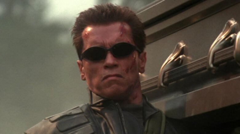 Uma captura de tela do Terminator de Arnold Schwarzenegger de Terminator 3, usando óculos escuros e segurando um caixão ornamentado em seu ombro