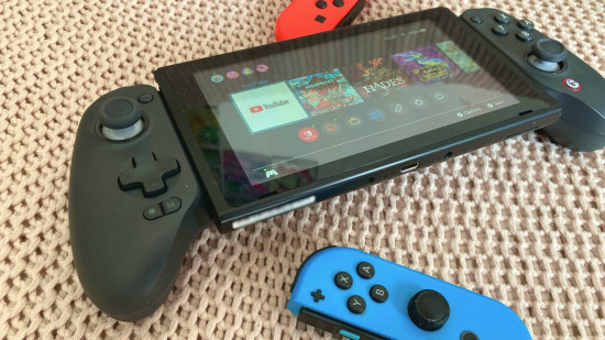 Controle Bluetooth GameSir G8+: O G8+ segurando um Switch na tela inicial, deitado em um cobertor rosa e fotografado de um ângulo leve. Joy-Cons vermelho e azul ficam ao lado do console