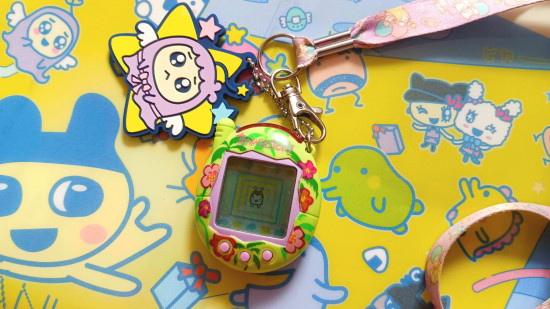 Tamagotchi revival: Um Tamagotchi Connection V3 verde e tropical deitado em um arquivo transparente com tema Tamagotchi. Ele tem um cordão rosa Little Twin Stars preso a ele, bem como um chaveiro de tira de borracha Weeptchi x Little Twin Stars